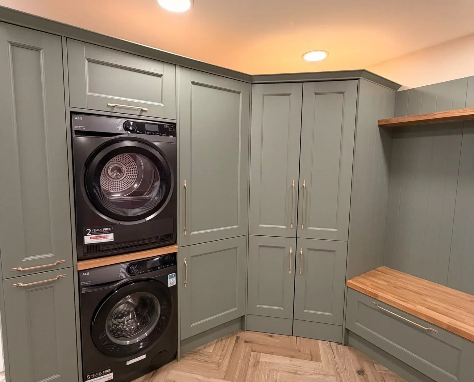 Washing machine and tumble dryer stacked in a sage green boot room with herringbone flooring